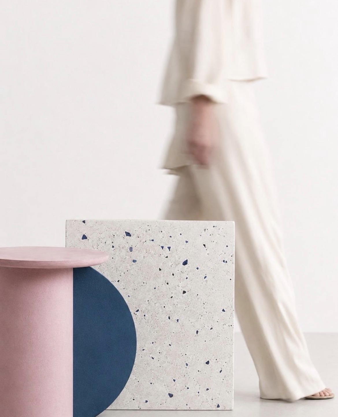 A pink cylindrical side table beside a stone-textured cube table with a navy inset, illustrating a range of custom colour and material options, a blurred figure walking past in the background.